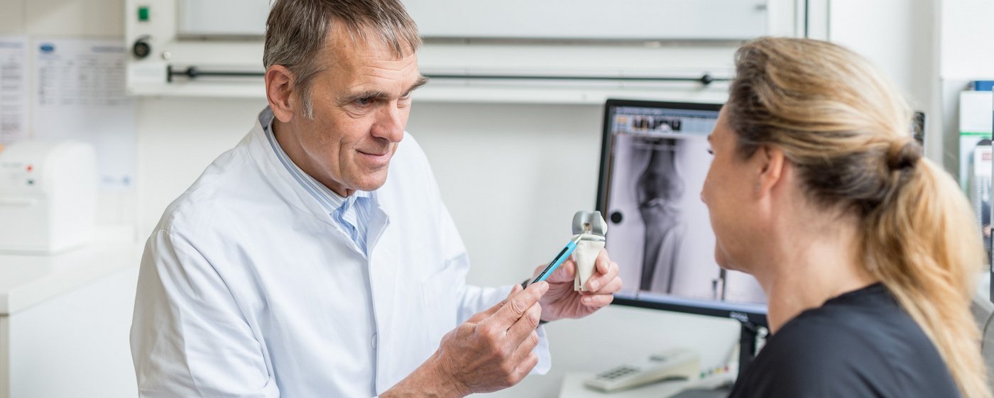 Ein Arzt erklärt einer Patientin die Funktionsweise eines Gelenks. Im Hintergrund ein Röntgenbild