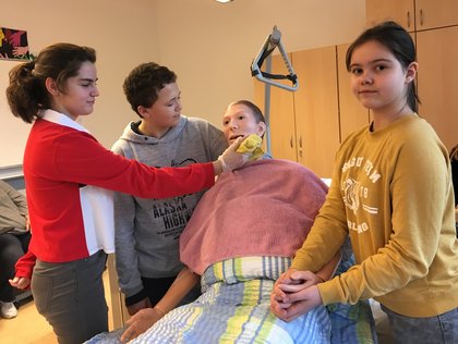 Drei Grundschüler üben die Pflege an einer Trainingspuppe im Bett und gucken begeistert