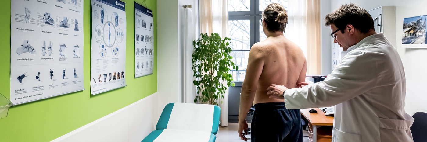 Ein Arzt überprüft die Hüfte eines Patienten, im Hintergrund hängen Poster zum Bewegungsapparat