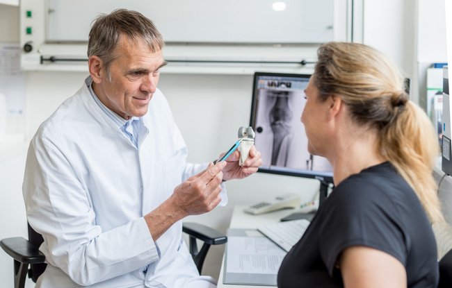 Ein Arzt erklärt einer Patientin die Funktionsweise eines Gelenks. Im Hintergrund ein Röntgenbild