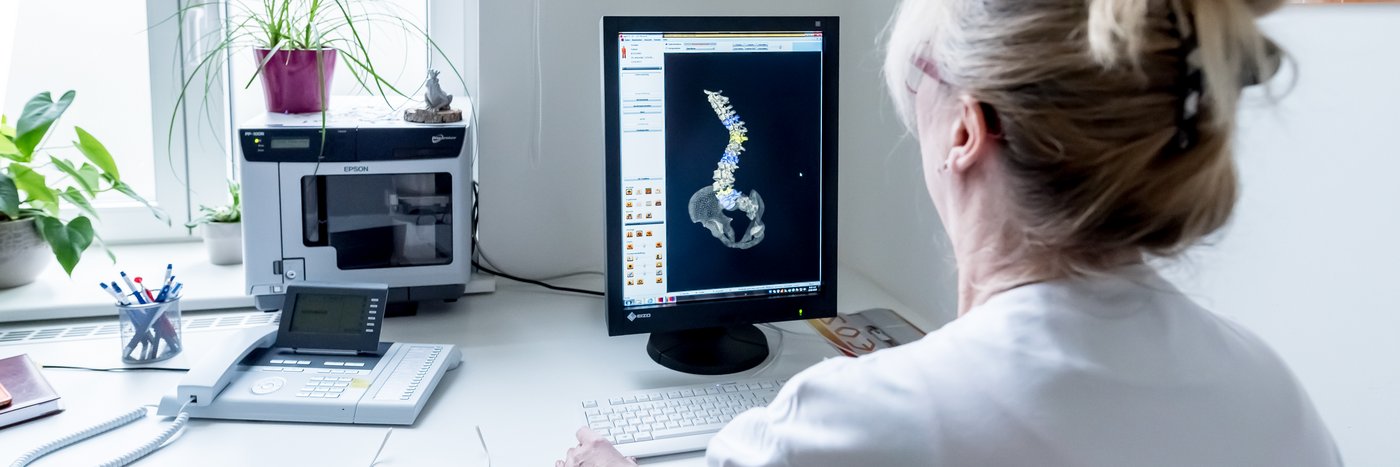 Eine Ärztin schaut sich den Verlauf der Wirbelsäule auf einem Monitor  an, Neben ihr Akten und eine Pflanze