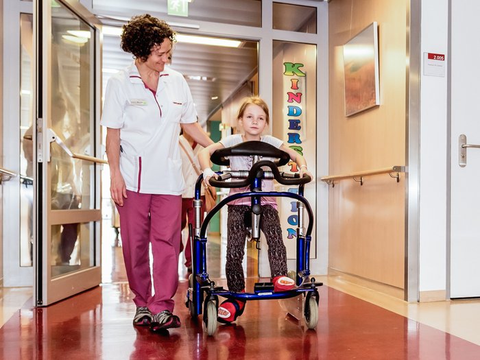 Ein Mädchen mit Bewegungshilfe wird von einer Krankenschwester mit freundlicher Ausstrahlung begleitet.