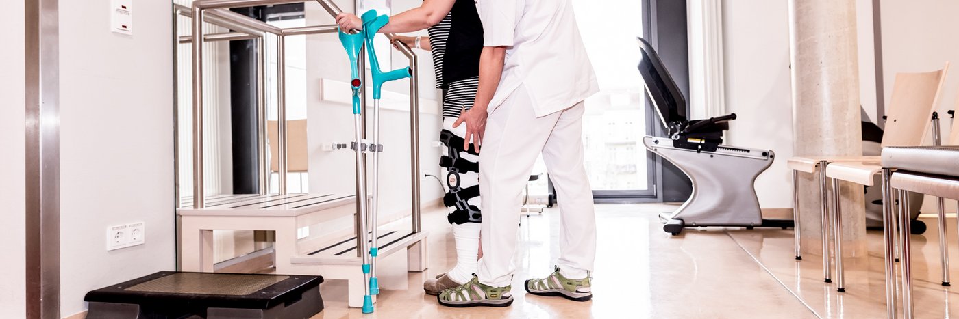 Ein Physiotherapeut überprüft die Beinschiene einer Patientin. Im Vordergrund steht eine Stütze  